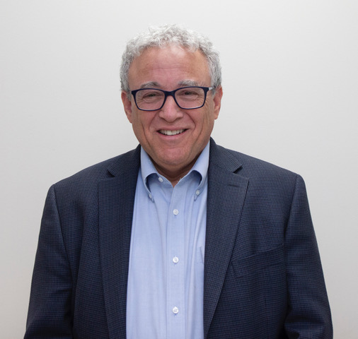 Howard Sitron headshot. Man wearing blue shirt and blazer, smiling wearing glasses and has short gray hair.