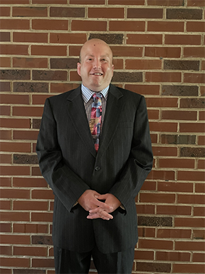 A white man with very short hair wearing a dark blue suit, and a stylish patterned shirt and tie smiles for the camera, standing in front of a brick wall.