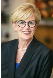 A smiling white woman with blonde hair and glasses, wearing a suit jacket.