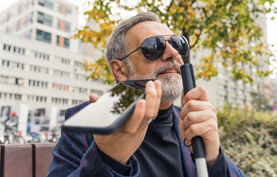 Blind middle-aged man with gray hair and beard wearing sunglasses holding cane and phone using text-to-speech function. 