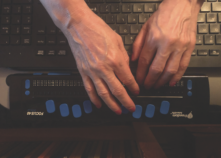 A view from above of the hands of a person with light skin reaching over a laptop keyboard to rest on a braille display. 