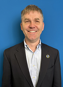 A smiling white man with blonde hair, wearing a black jacket and light blue shirt.