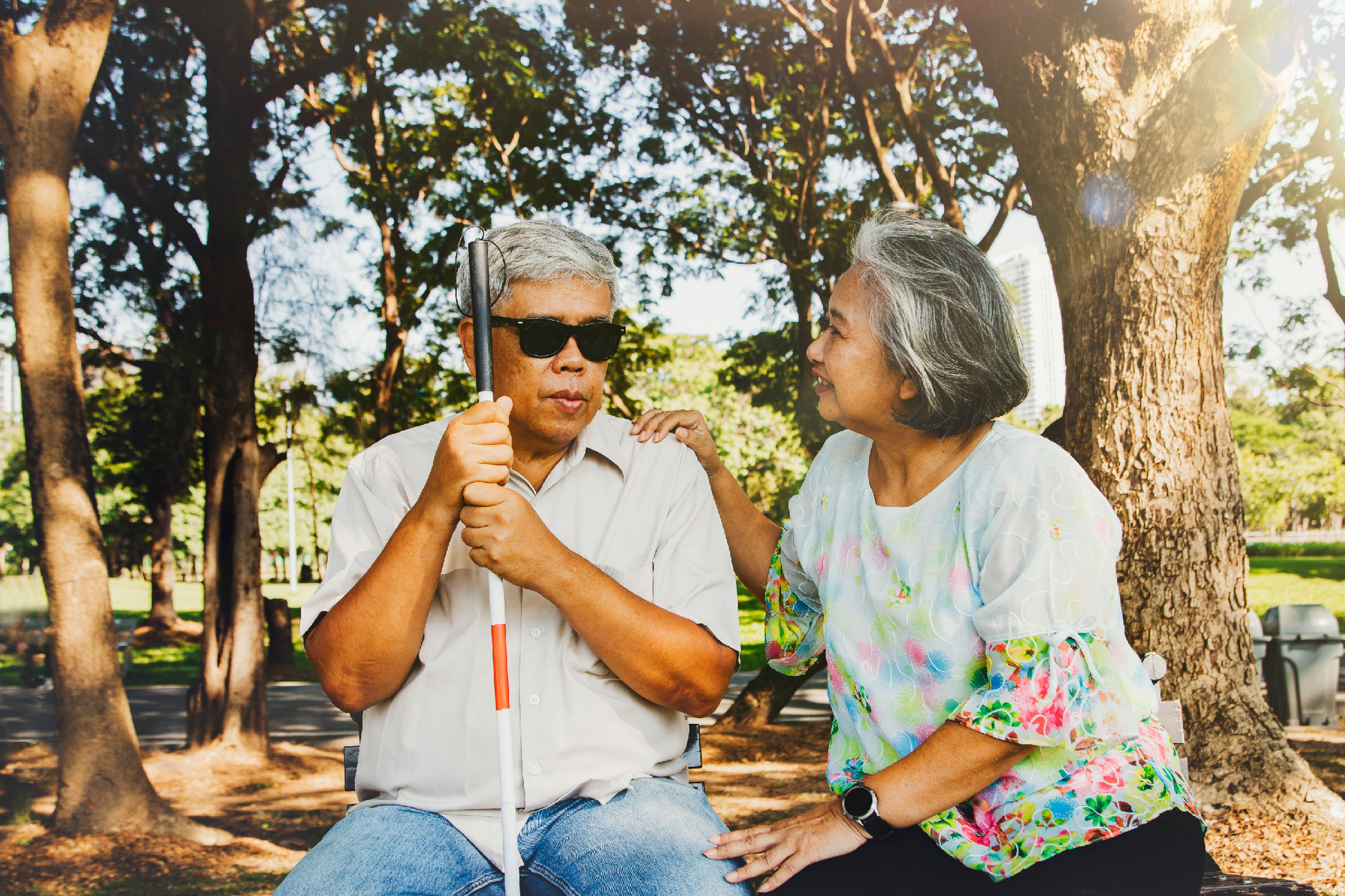 A person sits on a bench outside holding a white cane, and the person sitting next to them looks and smiles as they rest their hand on their shoulder