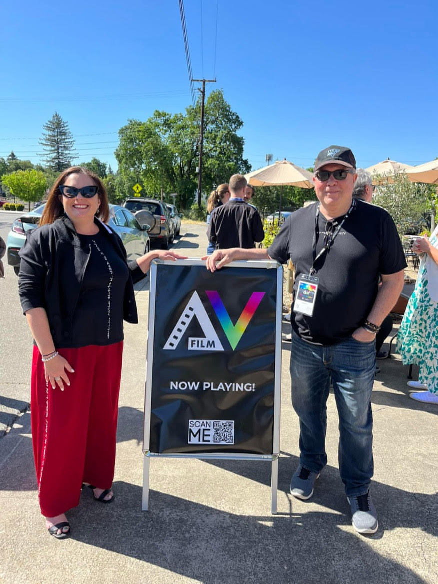 From left to right: Melody Goodspeed and Bill Sarine. They stand next to a sign that reads ''AV Film, Now Playing!"