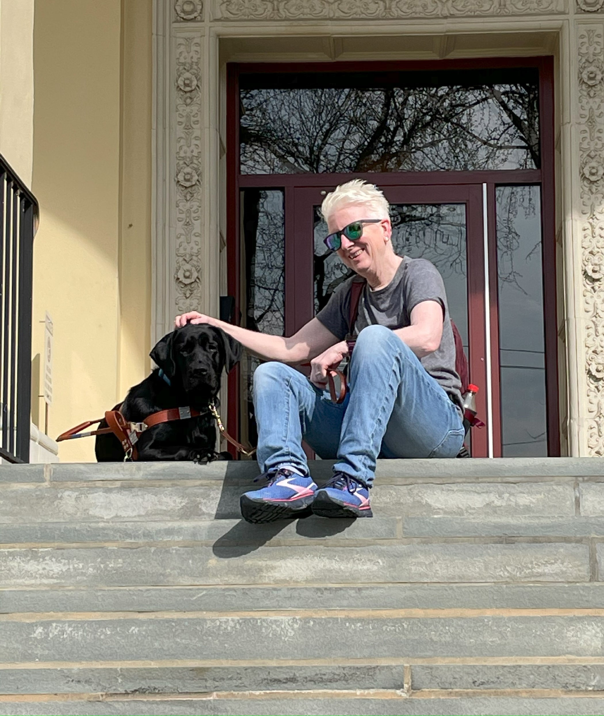 Pamela McGonigle sits at the top of a brick staircase next to her guide dog Mary. McGonigle smiles and pets Mary on her head.