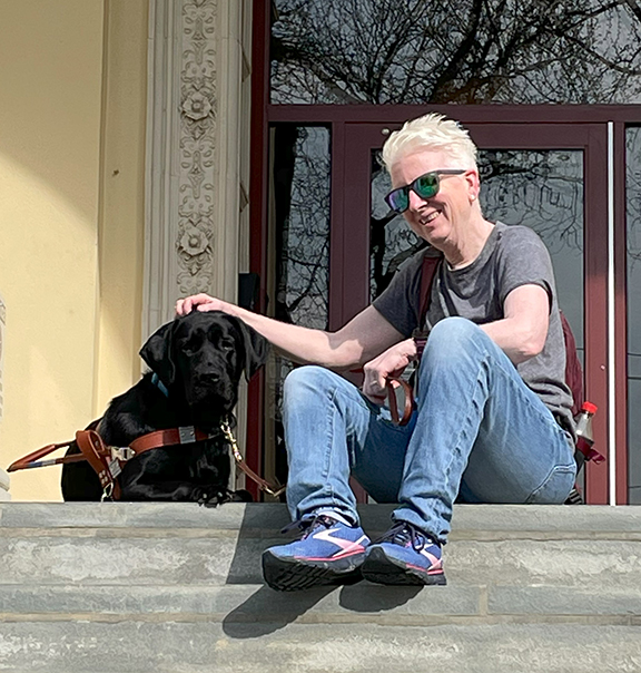 Pam, a smiling woman with short, platinum blonde hair, sits on building steps with her guide dog. 