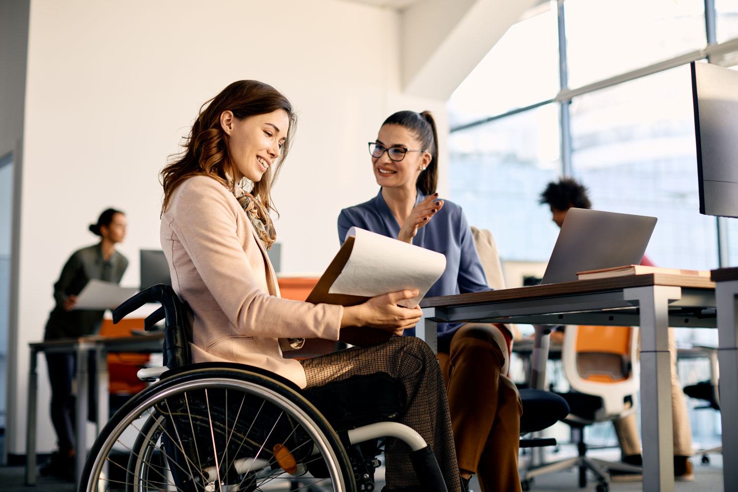 A businesswoman in a wheelchair going through reports while working with a female coworker in the office.