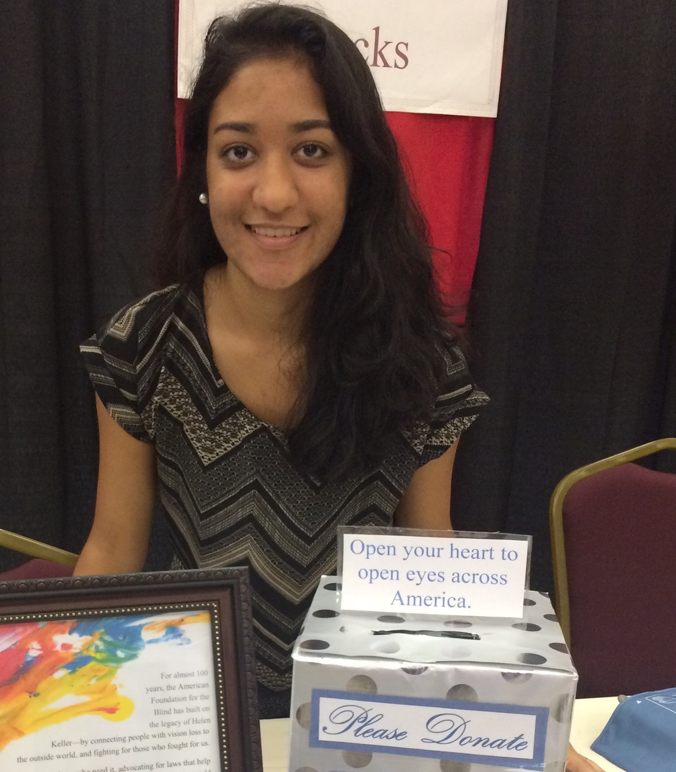 Poonam Agrawal sitting by a box that reads Please Donate: Open your heart to open eyes across America