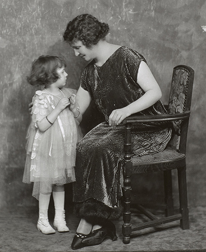 Helen Keller is seated in three-quarter profile, possibly in a photographer's studio. She is leaning over and has her arm around the back of a young girl who is standing next to her. Keller is wearing a loose, shimmering dress that is dark and has a boat neck. The child's dress is light-colored and made of layers of a delicate organza-type fabric. The photograph was taken in the 1920s.