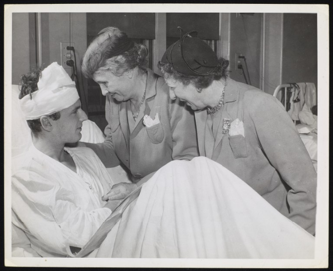 Taken during Helen Keller's tour of U. S. Military Hospitals. Keller and Polly Thomson are photographed in a military hospital ward, possibly in Pennsylvania? They stand on the far side of a bed where a veteran is sitting up. He has a bandage around his head and is bending his knees. The soldier is seen across the foreground of the image at a diagonal. The head of the bed is on the left-hand side of the image. A bedsheet covers him. The women are leaning towards him. Keller is nearest to his head and her right arm touches his back. The women wear very similar light-color jackets with a small pocket with a handkerchief sticking up. They both wear necklaces and hats. A metal-frame bed is visible in the background.