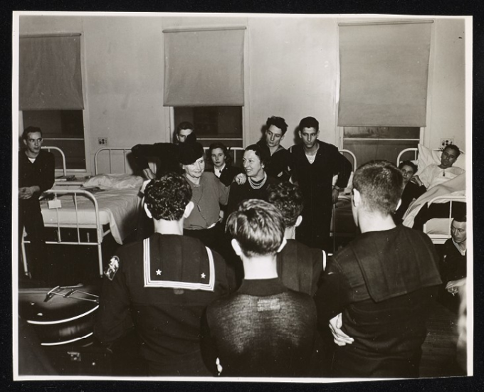 Taken during Helen Keller's tour of U. S. Military Hospitals. Keller and Polly Thomson stand in a ward at U. S. Naval General Hospital, Bainbridge, Maryland. Keller and Thomson stand in the aisle of a ward. Behind them is a row of metal-frame beds lined up against a wall with three windows and pull-down blinds. Servicemen and a servicewoman are sitting and leaning against the beds, A patient is in one of the beds. All of them are looking from behind at Keller and Thomson. The backs of four sailors fills the foreground of the image as they watch Keller and Thomson. A guitar can be seen lying flat on the left-hand side of the photograph. Keller's left hand is on Thomson's right shoulder.