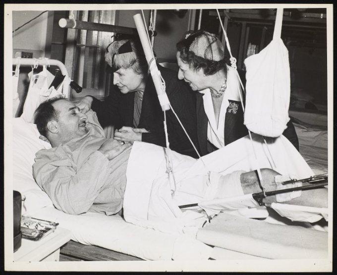 Taken during Helen Keller's tour of U. S. Military Hospitals. Keller and Polly Thomson are in a ward at Lovell General Hospital, Ayer, Massachusetts. A veteran is pictured lying in a metal-frame bed. The bed cuts across the foreground of the image at a diagonal, with the head of the bed on the left-hand side of the image. The soldier is wearing pyjamas, a bedsheet covers his waist and left leg. His right leg is visible from just above the knee to his calf. It is suspended in a sling from a contraption above him. Thomson and Keller who stand on the far side of the bed are bending over towards him. Keller is nearest to his head, her right hand is on his pillow. Keller is possibly holding the veteran's right hand in her left hand and Thomson hold's Keller's left wrist. The women wear hats and dark pinstripe jackets