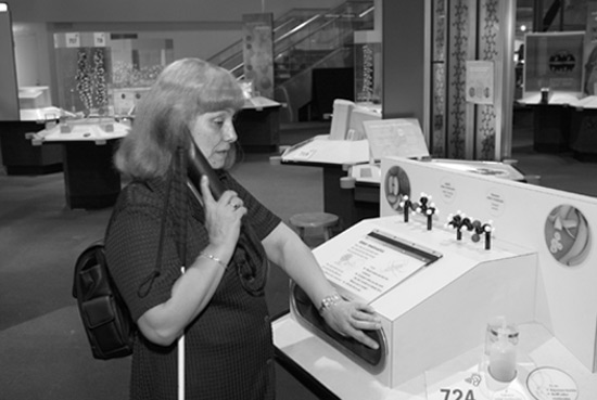 Figure 1: Photograph of woman with a long cane listening to audio tour phone and reaching into a box under a hands-on display showing a model of molecules and photographs of fruit.