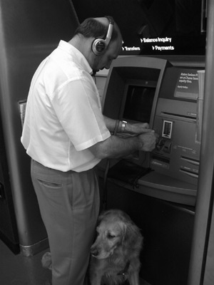 Figure 1: Photograph of a man wearing a headset in front of a Chase e-ATM feeling to the side of the keypad for the headset jack, while a dog guide waits.