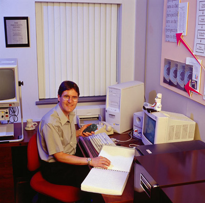 Photo of Peter Torpey at his desk with a computer, braille display, remote control, braille book, and Dilbert doll.