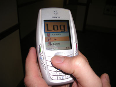 A Nokia cell phone in a user's hand. The screen displays the magnification window at the top, showing the word "Log." The rest of the screen shows "Contacts," "Log," and "Gallery," with icons for each.