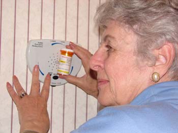 A gray haired woman holding a prescription bottle up to the ScripTalk device to have information read aloud to her.