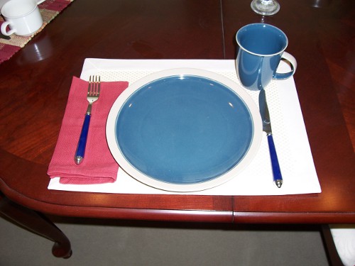 High contrast place setting: dark table with white place mat, blue plate and cup, and blue-handle utensils, red napkin.