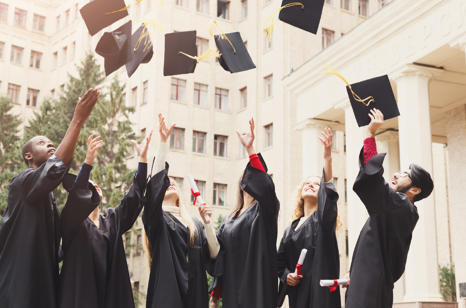 A group of students in graduation gowns throw their caps in the air.